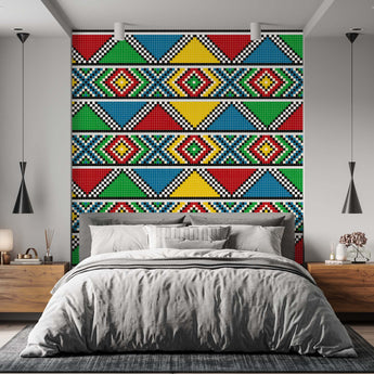 On a wall is a Zulu inspired wallpaper with a geometric repeat pattern of triangles and diamond shapes. Colours red, green, blue, black and white. Setting is a bedroom with a bed and wooden pedestals